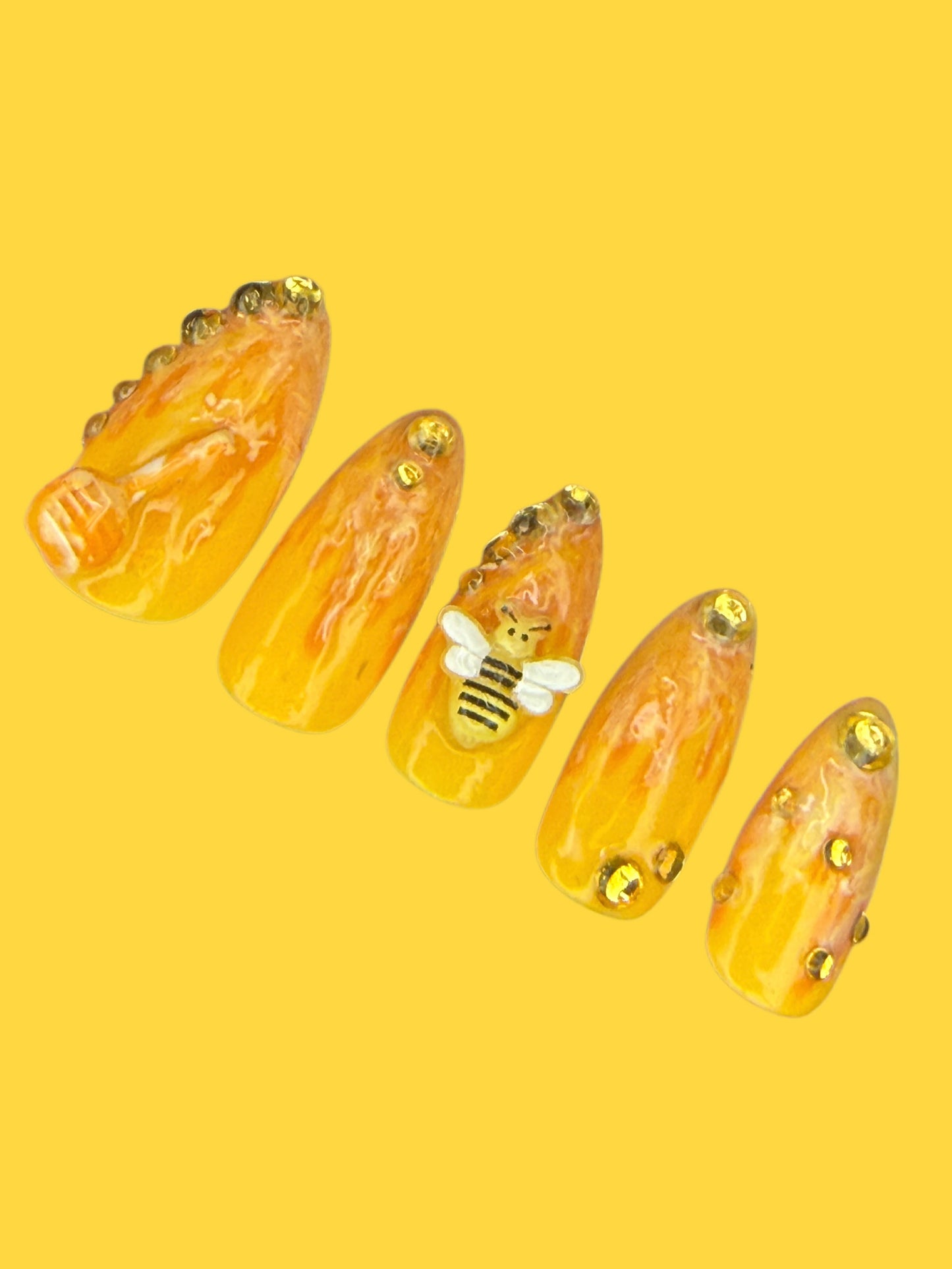 Five almond-shaped press-on nails with a honey drip pattern in translucent amber-yellow, decorated with gold rhinestones and a small white-and-black bee on one nail, set against a solid yellow background.