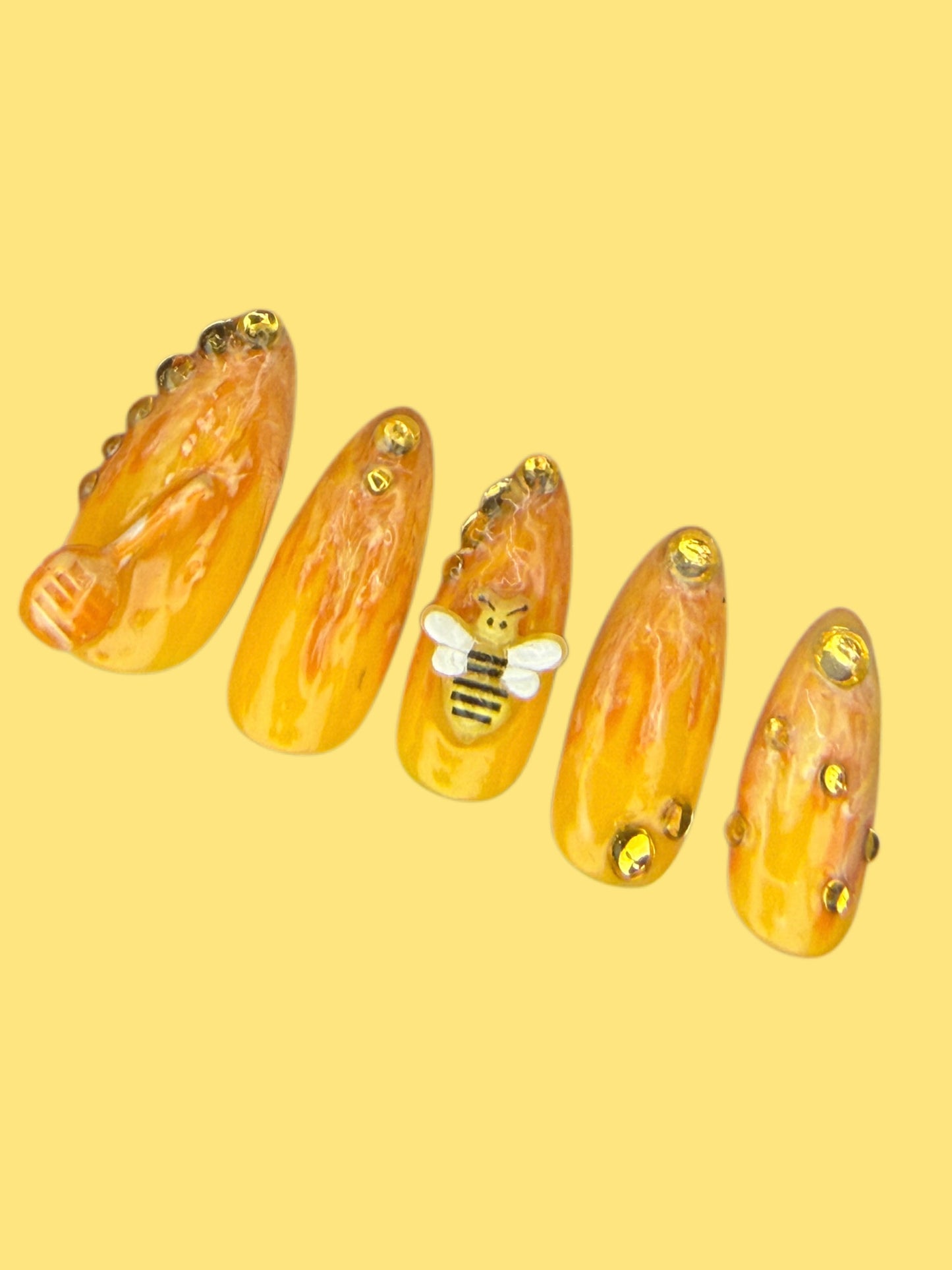 A set of five almond-shaped press-on nails with a honey drip pattern in warm amber and gold, decorated with small gold rhinestones and a central bee accent, displayed on a solid pale yellow background.